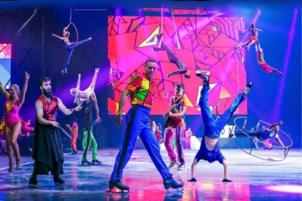 Ice skaters in colorful costumes perform as acrobats perform suspended in the background