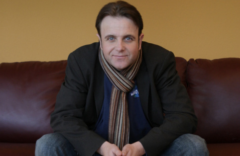 Man sitting on brown couch. He has short black hair and blue eyes. He is wearing a loose blazer with a blue t-shirt underneath. He has on a stripped scarf.