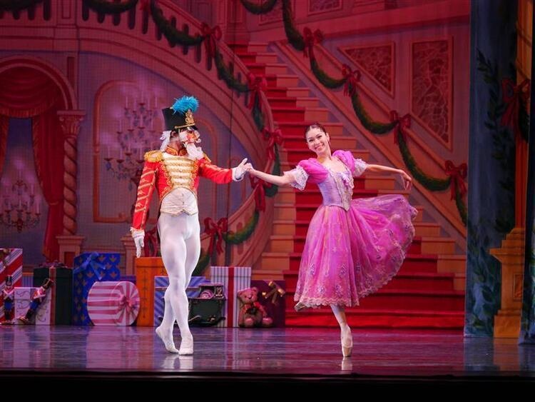 Two ballet dancers perform on stage in front of a festive backdrop with red stairs adn oversized holiday decorations. One dance is dressed as a toy soldier, and the other wears a bright pink dress, en pointe with one leg extended.