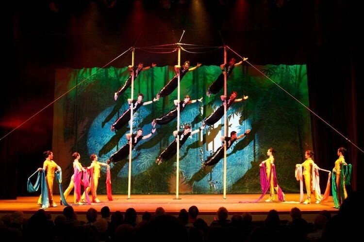 Performers in colorful costumes balance on poles and pose in front of a vibrant stage backdrop during a theater production.