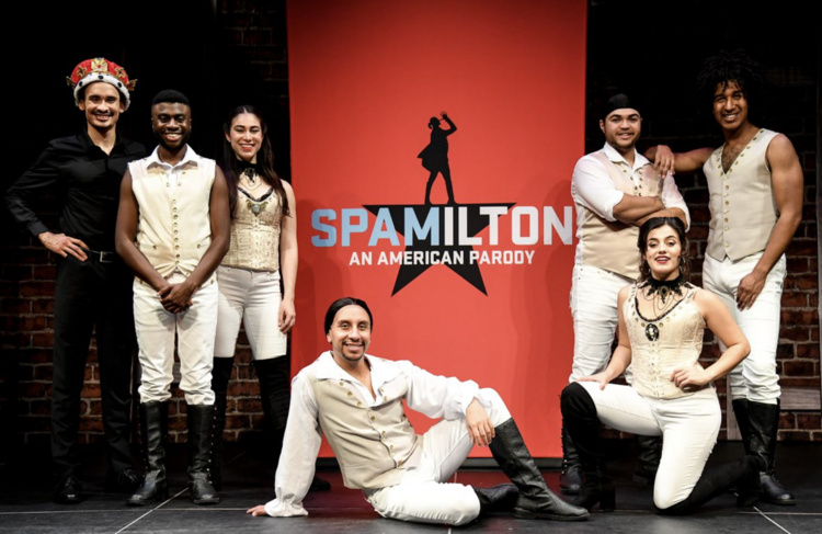Seven performers pose on stage in front of a red backdrop that reads "Spamilton: An American Parody," featuring a silhouette of a performer holding a microphone. The cast wears coordinated costumes reminiscent of 18th-century American revolutionary style, with cream-colored vests, white pants, and black boots. One performer wears a crown, suggesting a comedic portrayal of royalty. The group is smiling and arranged in a friendly, theatrical pose, emphasizing the show's lighthearted and satirical tone.
