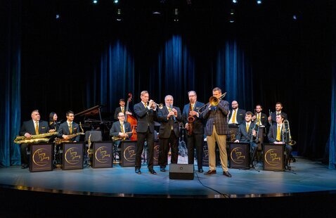 14 people on a stage. 10 are sitting with instruments the other four are standing playing their instruments. They are all dressed formal attire. There is one woman, the other 13 are men.  