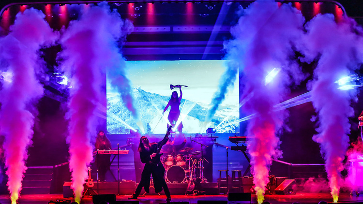 A dramatic concert scene, featuring a silhouetted performer in the center of the stage with a microphone. There are two other visible performers, one with an instrument raised above their head in front of a digital screen with a mountain scape. Colored smoke cannons and lights make a vibrant pink, purple, and yellow hue. The person in the center is playing a guitar and another is playing a keyboard.