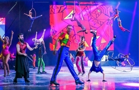 performers in colorful costumes dance and do acrobatics on ice stage with a bright pink geometric backdrop.