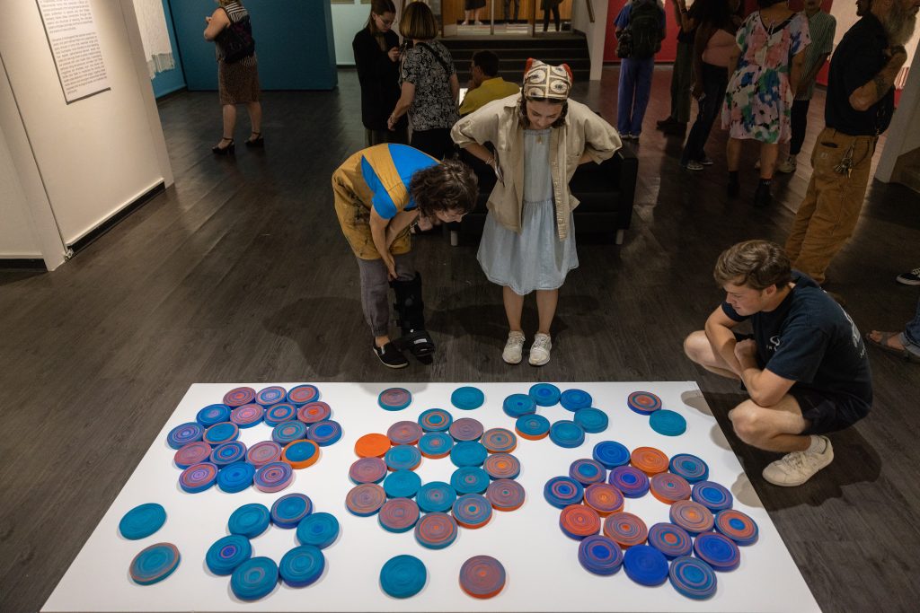 Three gallery visitors bend over and kneel to view an art installed on the gallery floor