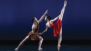 A male and female ballet dancer perform on stage. The male wears a brown costume, extending one arm, while the female in red gracefully lifts her leg high.
