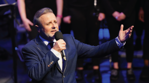 Michael passionately sings into a microphone on stage, extending one arm. Blurry audience members in black can be seen in the background.
