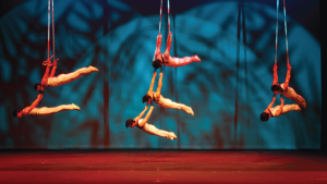 Aerial performers in matching outfits gracefully hang upside-down on red ribbons against a blue-lit abstract backdrop, conveying a sense of elegance and strength.