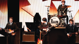 Four musicians in suits perform on stage, resembling a famous 1960s band. One plays drums, while the others play guitar and sing. Vintage vibe, energetic mood.