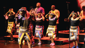 erforms on stage, wearing colorful traditional attire with striped patterns. The lead singer holds a microphone passionately singing.