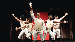 Five performers in beige vests and pants energetically pose on stage, arms outstretched. The center holds a doll aloft, conveying excitement and celebration.