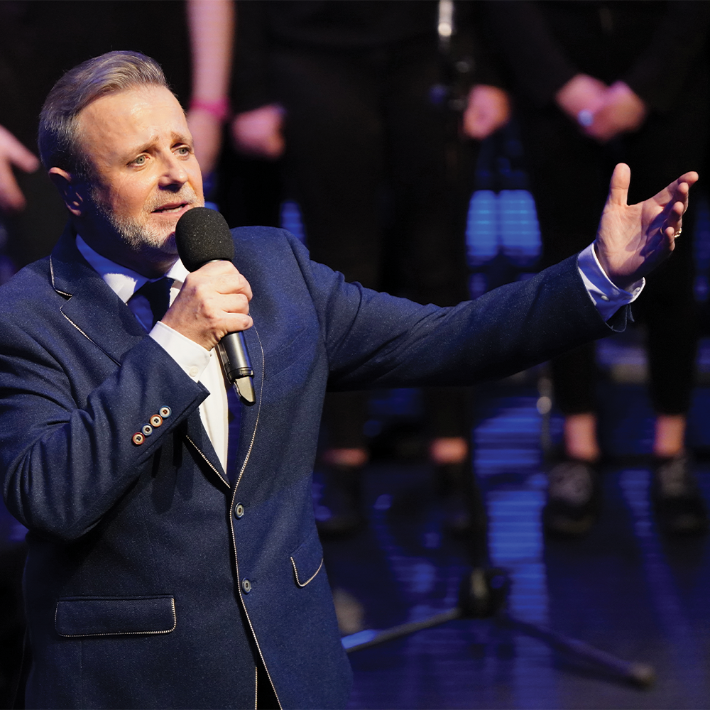 A man in a blue suit passionately sings into a microphone on stage. He gestures with one hand, conveying emotion. A choir stands blurred in the background.