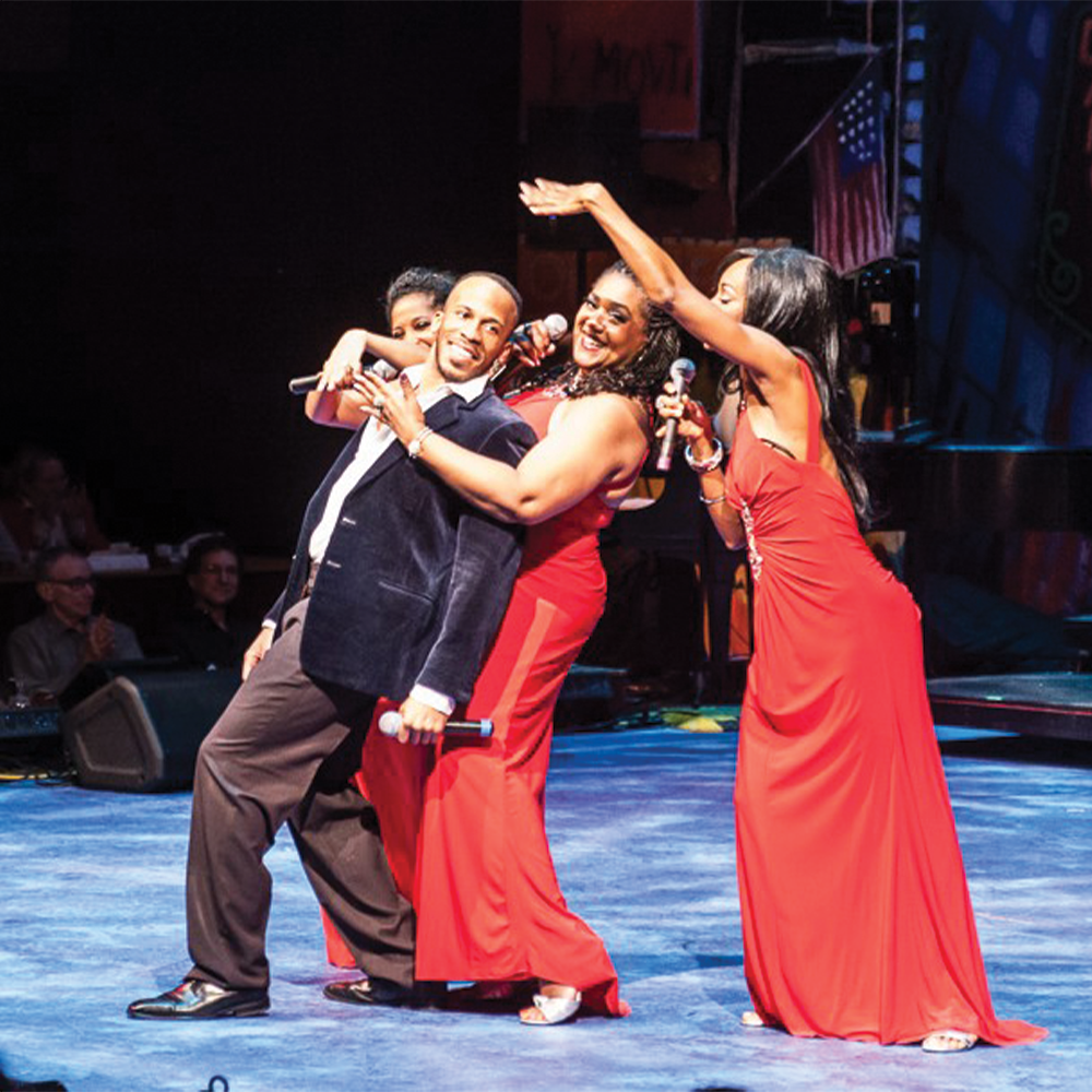 Four performers on stage, one man and three women, all wearing elegant attire. The women embrace the smiling man with microphones in hand.