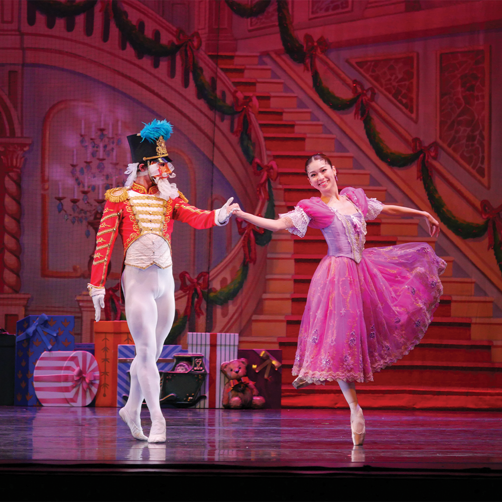 Ballet dancers perform on stage; one in a vibrant soldier's costume, the other in a flowing pink dress. Festive backdrop with gifts and garlands.