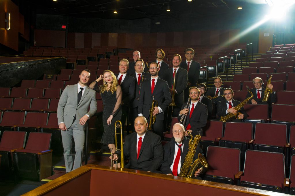 The Glenn Miller band sit in a Theatre auditorium while holding their instruments. 