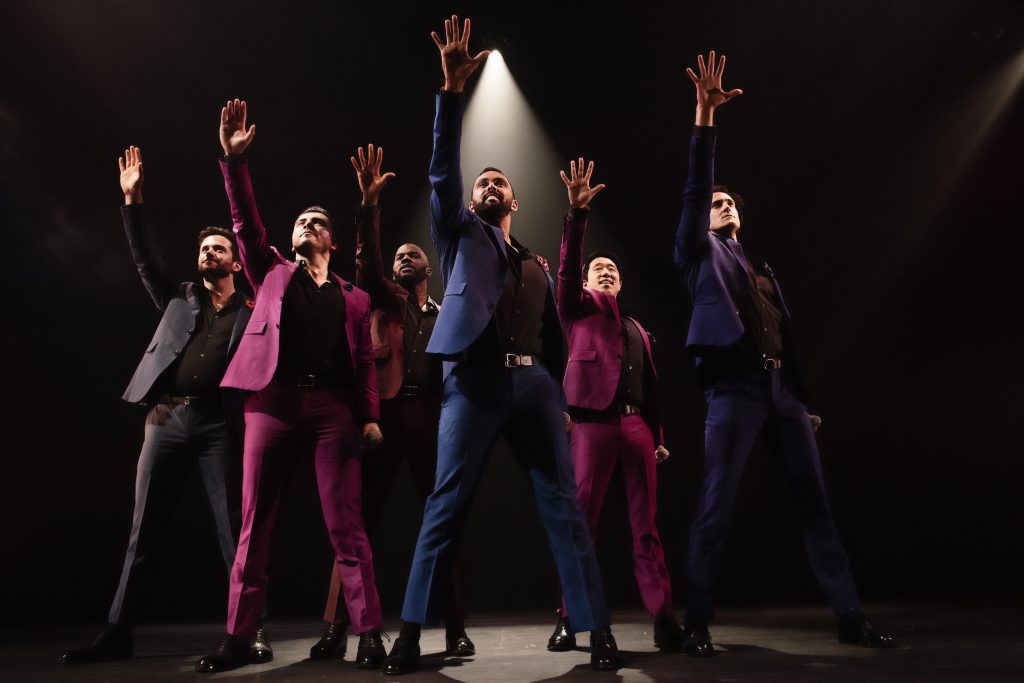 The Broadway Boys sing while standing in the middle of the stage, each with one arm raised up toward a single overhead light.