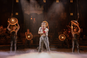 Performer on stage under dramatic lighting, surrounded by other performers. The scene conveys intensity and emotion in a theatrical setting.