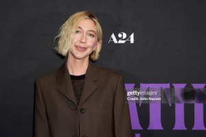 Heidi Gardner, with short, wavy hair, smiles subtly, wearing a brown blazer against a black background. The tone is calm and confident.