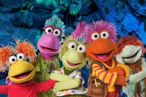 Five colorful puppet characters with vibrant hair and expressive faces cheerfully pose against a cave-like background, conveying a fun and friendly atmosphere.