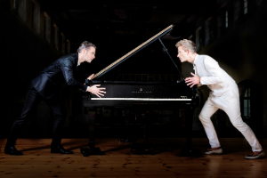 Two men, one in a black suit and the other in white, face each other intensely over a grand piano in a dimly lit room, creating a dramatic atmosphere.