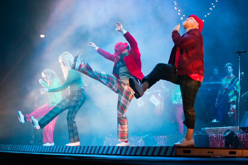 Three performers dance on stage wearing festive Santa caps and doing high kicks.