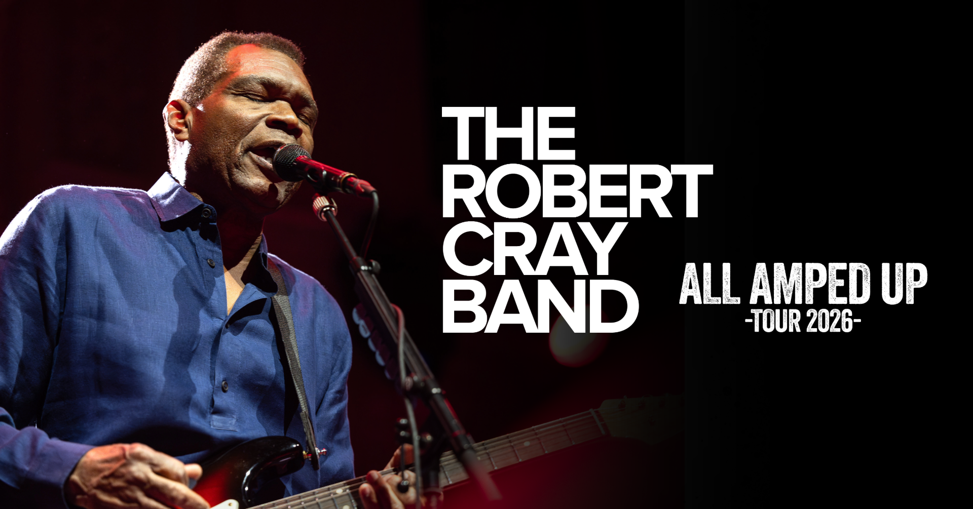 Robert Cray sings into a microphone while holding a guitar.