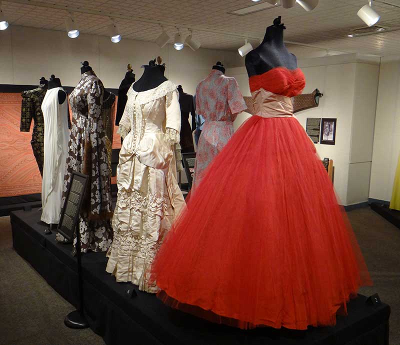Exhibit of elegant vintage dresses on mannequins, featuring a striking red gown with a full tulle skirt, set in a softly lit museum space.